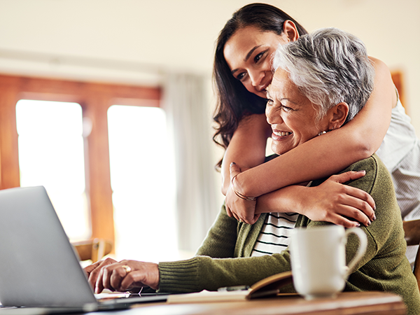 Older woman working on laptop while adult daughter gives hug from behind