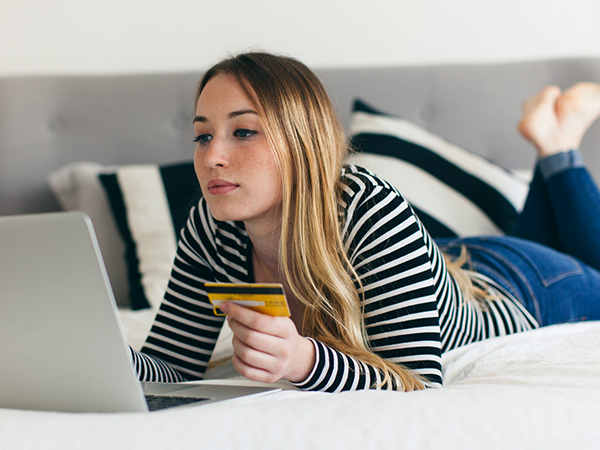 Woman lying on her bed looks at her computer while holding a credit card