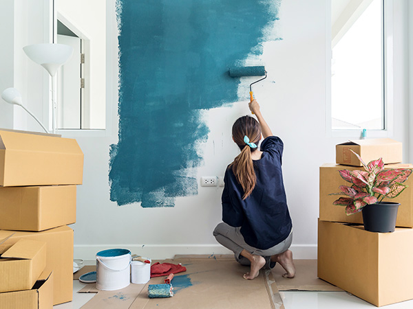 Woman paints walls with a roller with unpacked moving boxes surrounding her.