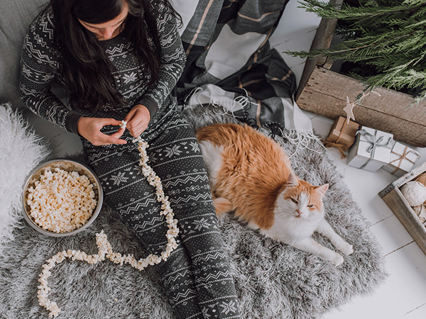Woman strings popcorn onto a holiday garland with cat resting beside her.